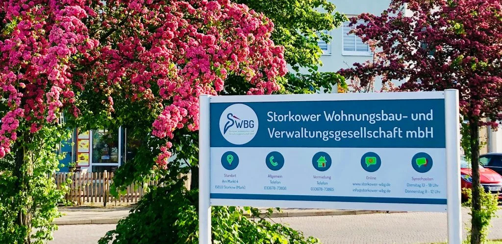 Firmenschild der Storkower WBG Am Markt 4, umrahmt von blühenden Rosen